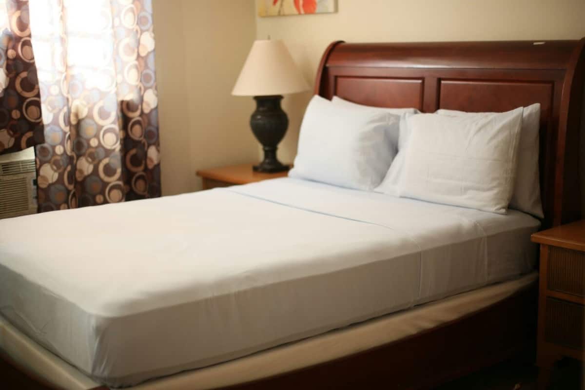 A neatly made bed with white linens and two pillows, a wooden headboard, a bedside lamp, and a patterned curtain.