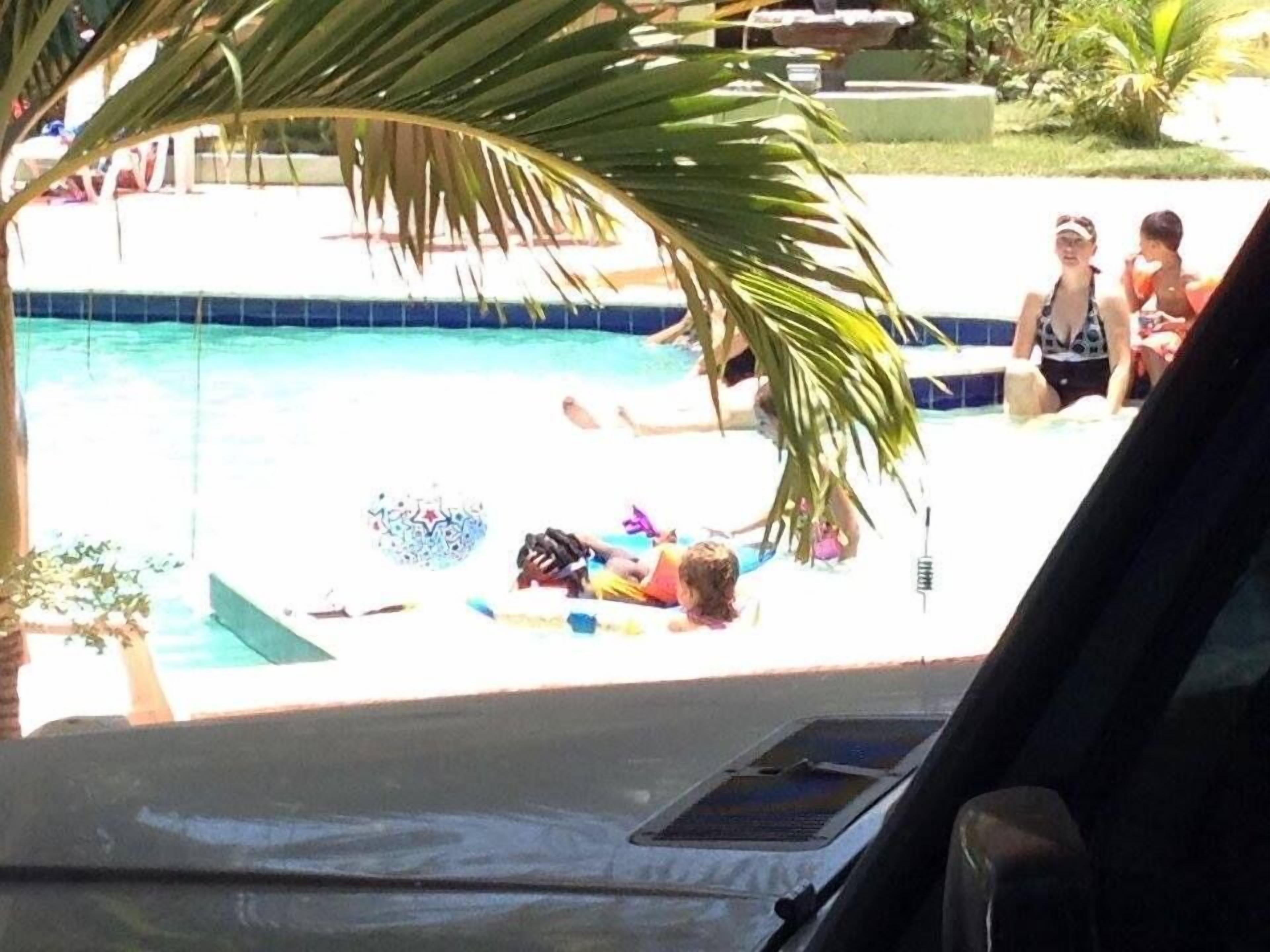 A swimming pool with a tiled deck, surrounded by palm trees and a few people.