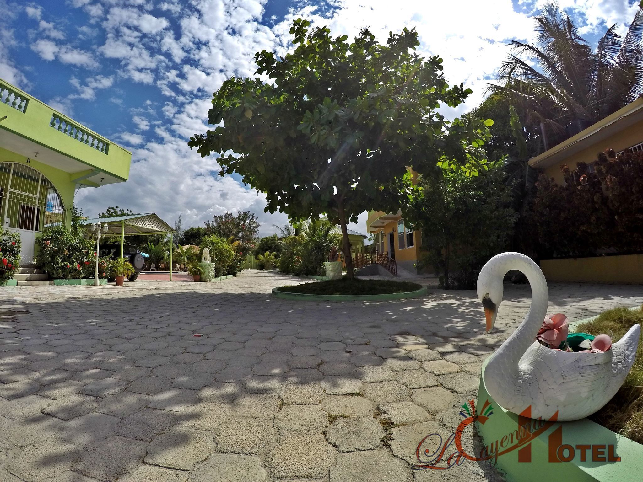 A courtyard with a swan sculpture, paved with hexagonal tiles, surrounded by greenery and a building with a green facade.