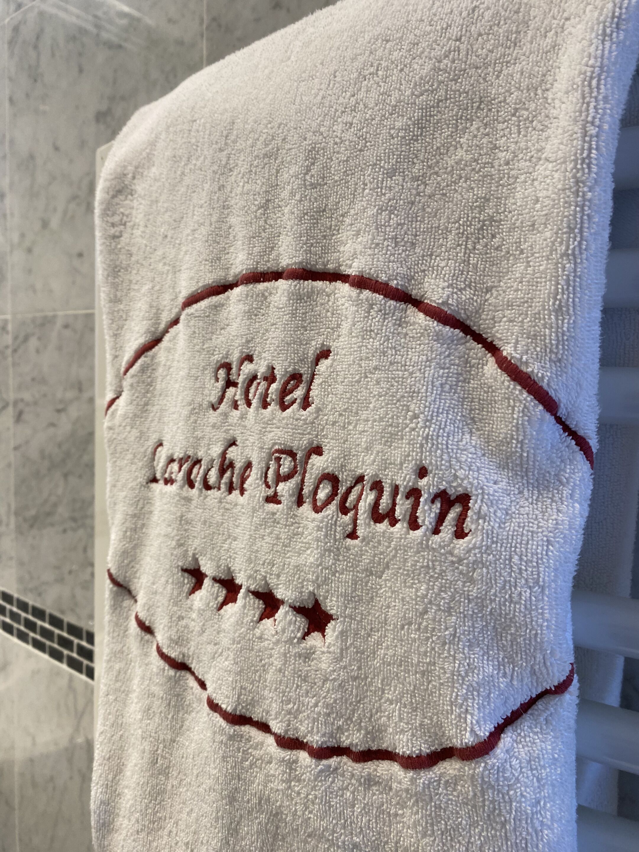 Une serviette blanche ornée d’un texte brodé indiquant « Hôtel La Roche Ploquin » et de motifs en forme d’étoiles.
