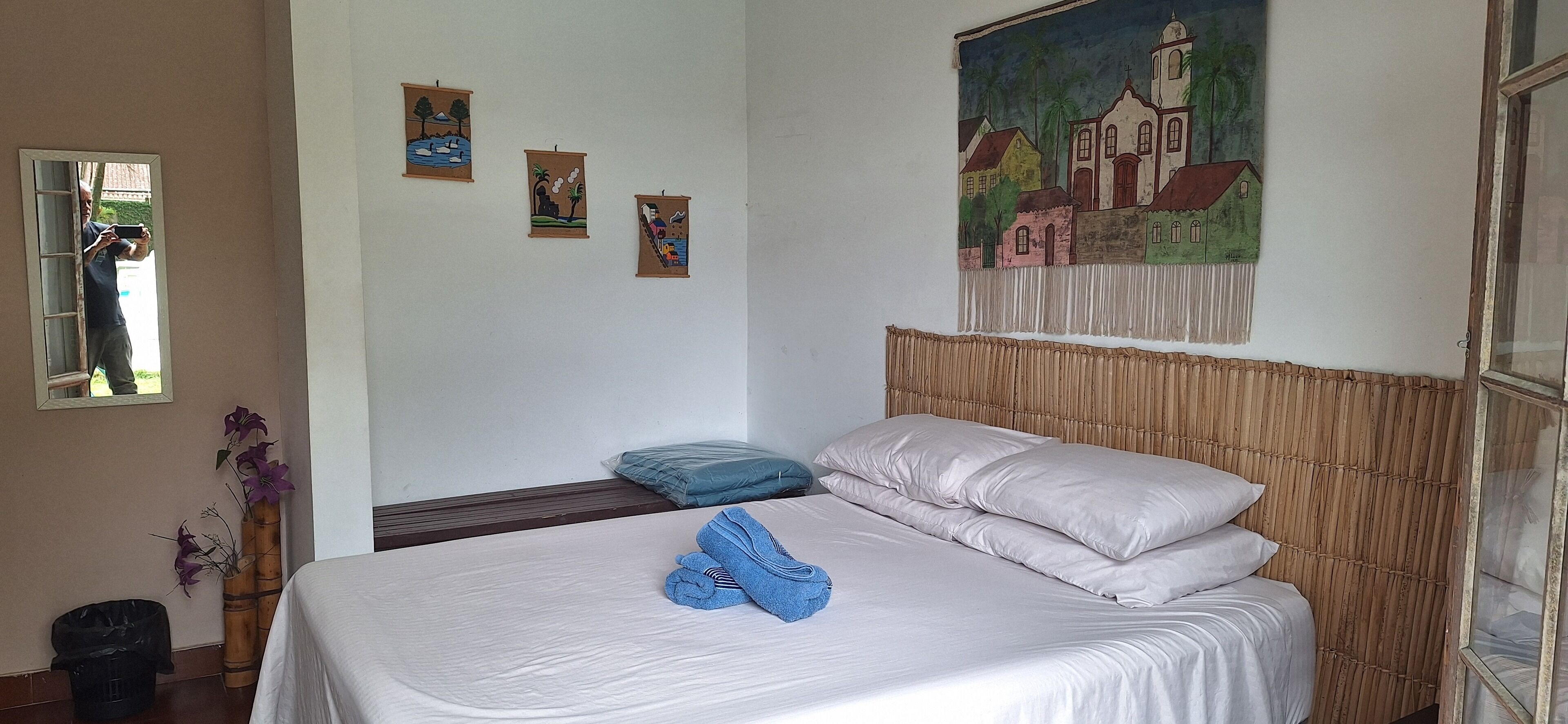 A neatly made bed with pillows and a folded towel, a mirror, and three small paintings on the wall.
