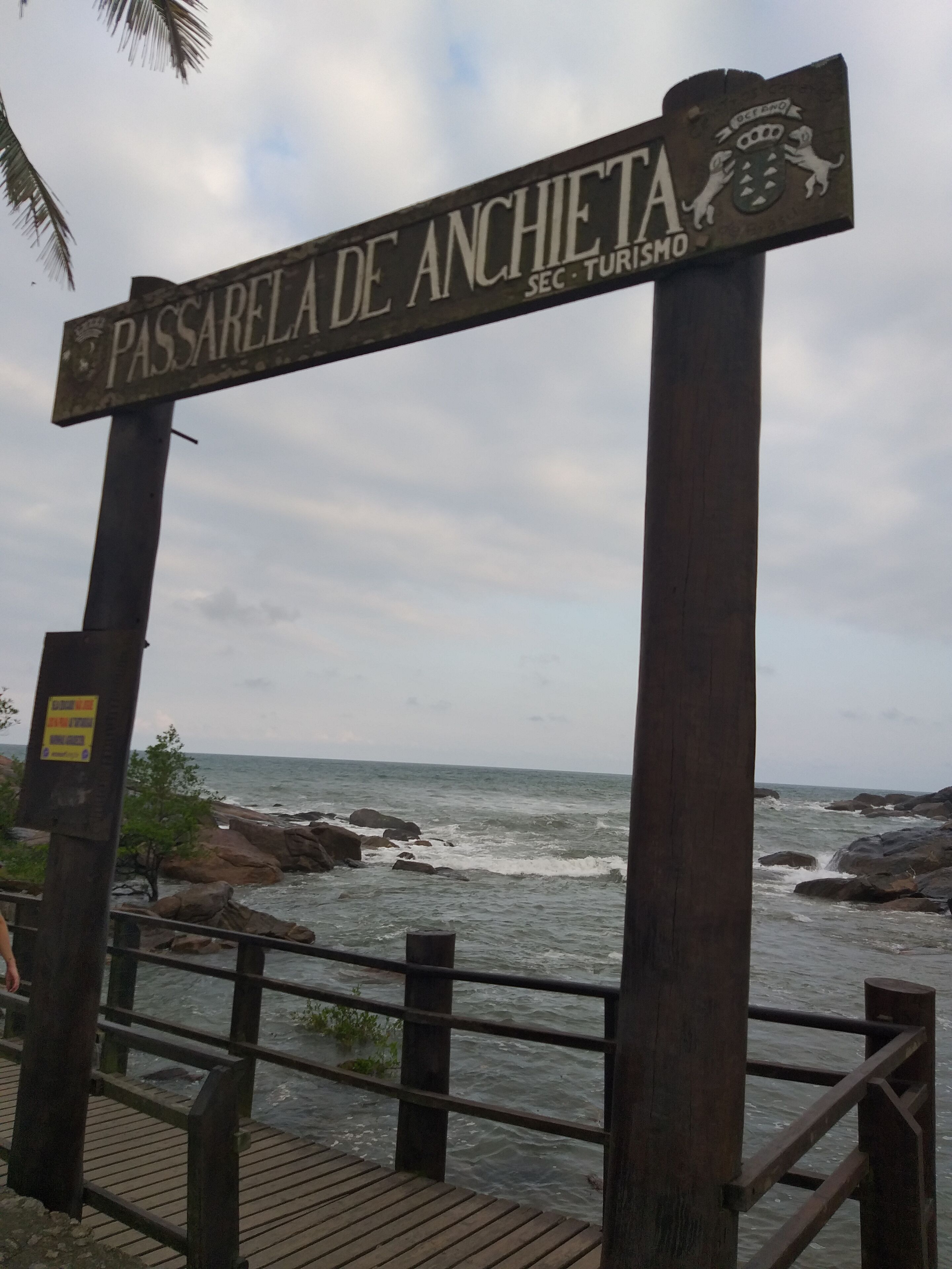 Un cartel con la inscripción 'Passarela de Anchieta' y 'SEC. TURISMO', con vistas a una costa rocosa.