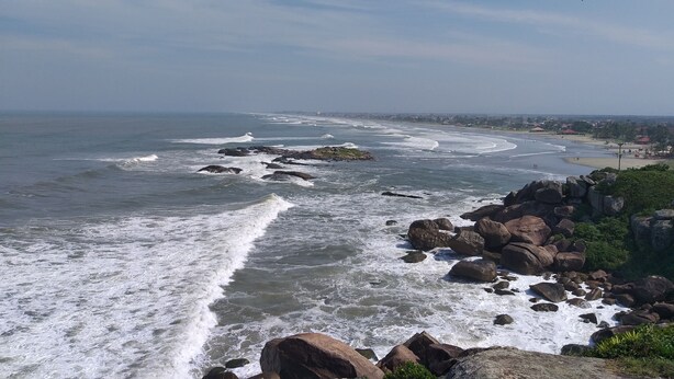 Vista costera con olas rompiendo contra rocas, una playa arenosa y un pueblo lejano.