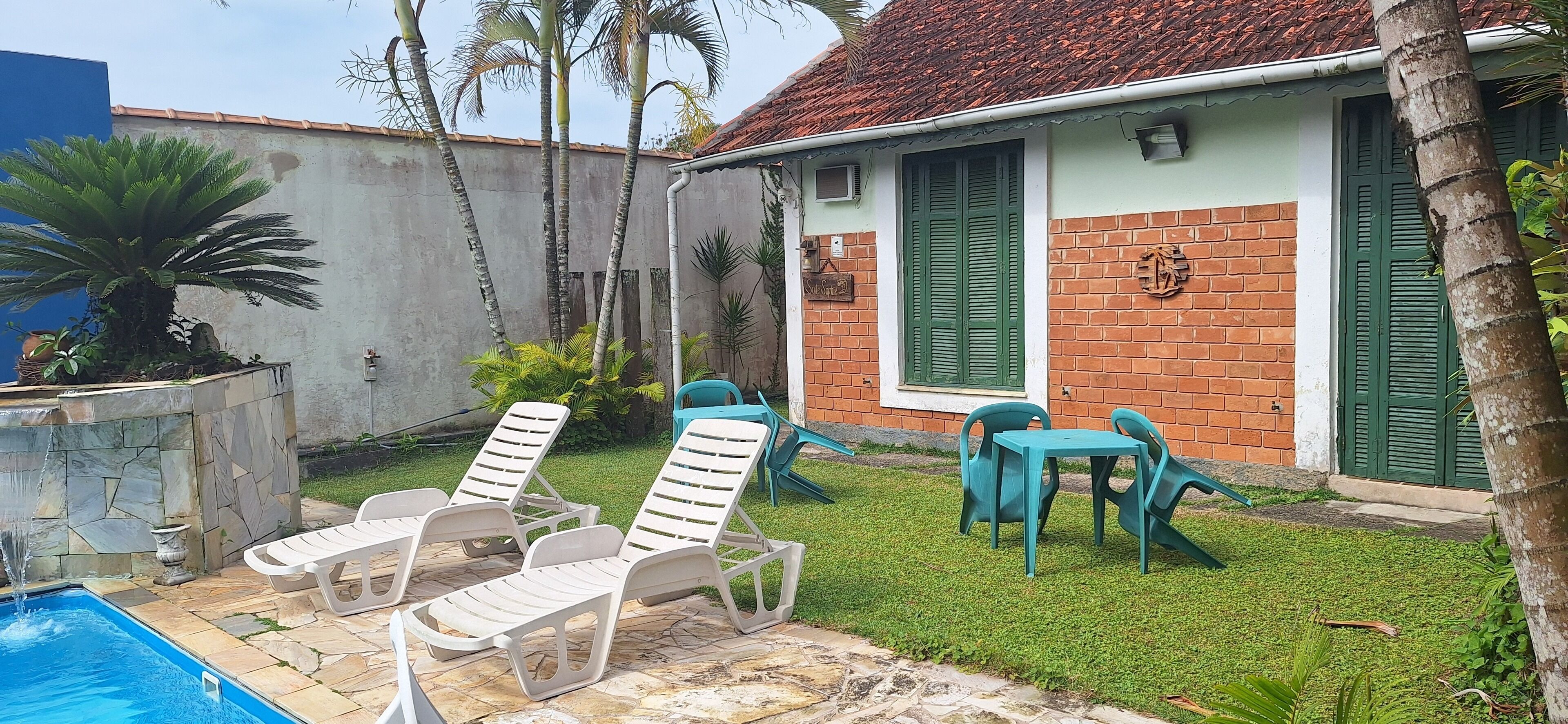 A pool area with lounge chairs and a table, surrounded by a small garden with palm trees and a brick building with green shutters.