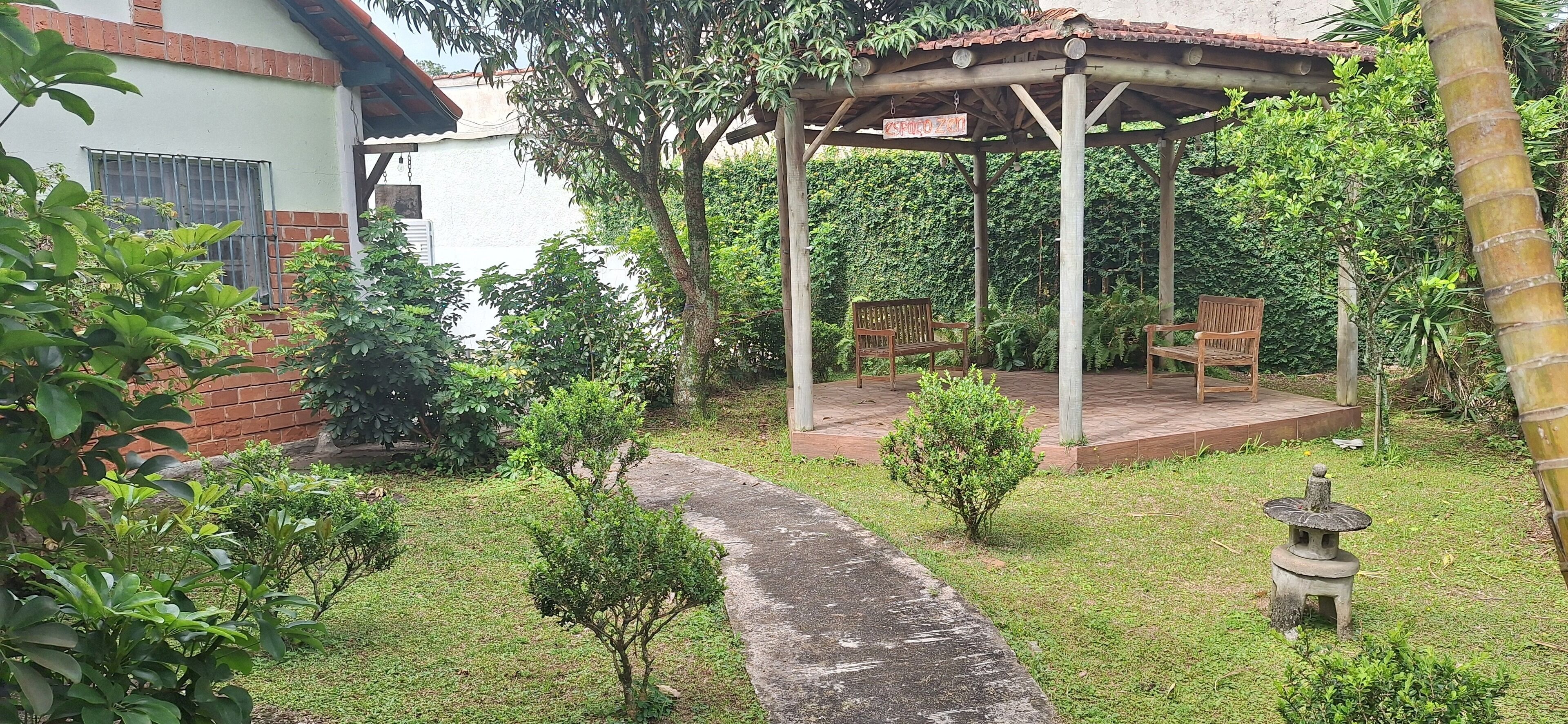 Uma área de estar coberta ao ar livre, com um banco de madeira e uma mesa pequena, rodeada por vegetação exuberante e um caminho de pedra.