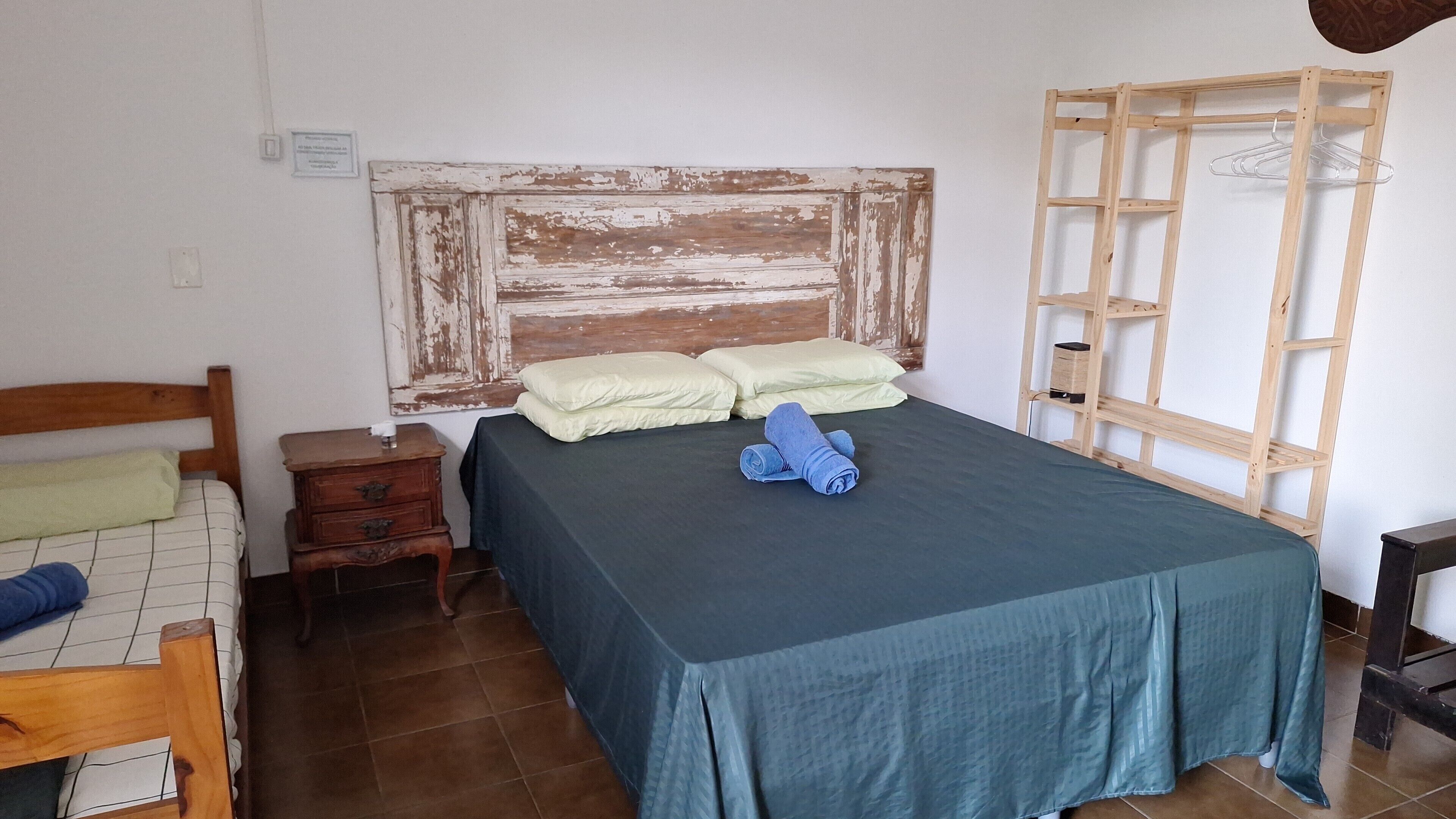 Un dormitorio con cabecera de madera, cama con colcha verde, dos almohadas, mesita de noche de madera con lámpara y una estantería de madera con perchero.