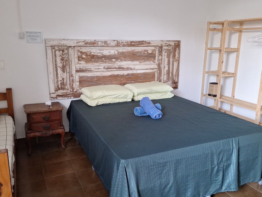 Un dormitorio con cabecera de madera, cama con colcha verde, dos almohadas, mesita de noche de madera con lámpara y una estantería de madera con perchero.