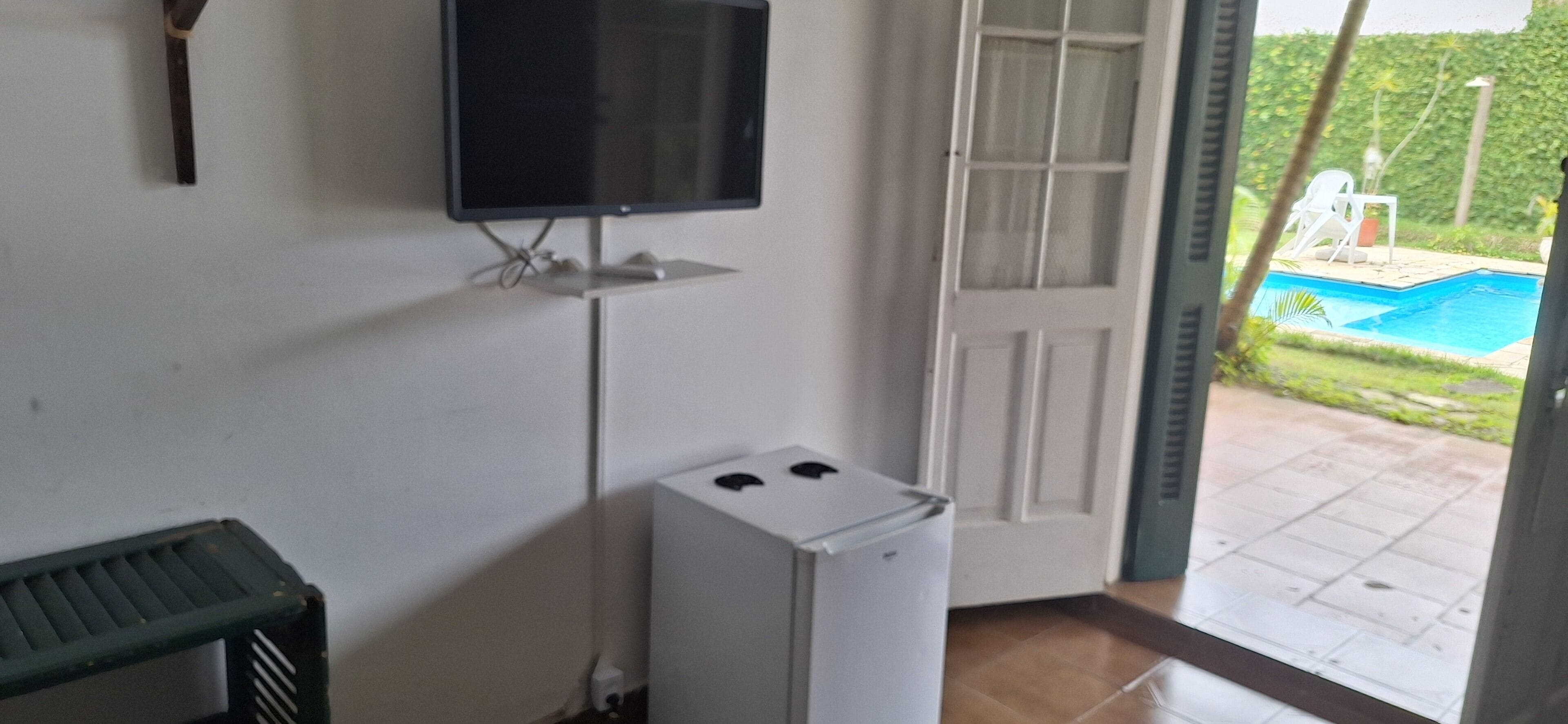 A room with a television mounted on the wall, a small white refrigerator, and a door leading to an outdoor area with a pool.
