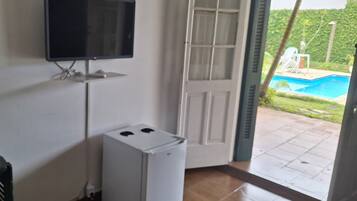 A room with a television mounted on the wall, a small white refrigerator, and a door leading to an outdoor area with a pool.