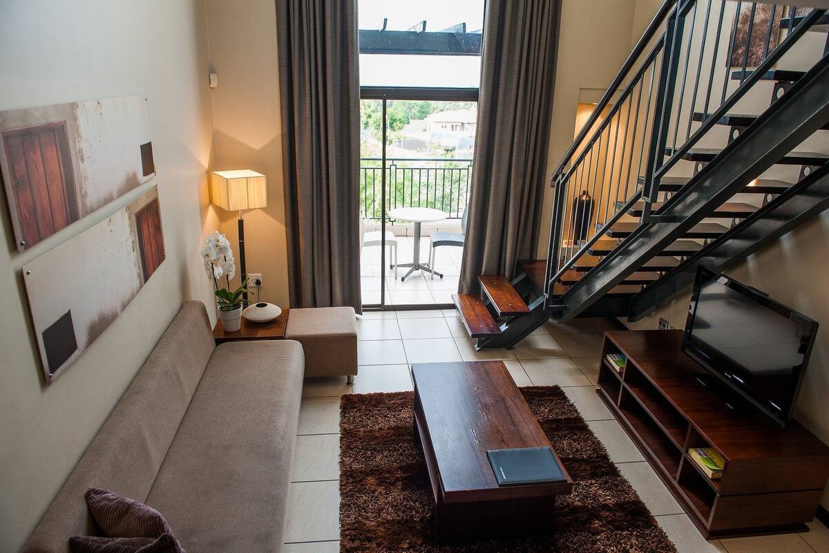 A living room with a sofa, a coffee table, a TV on a wooden stand, a lamp, and a view of the outside balcony.