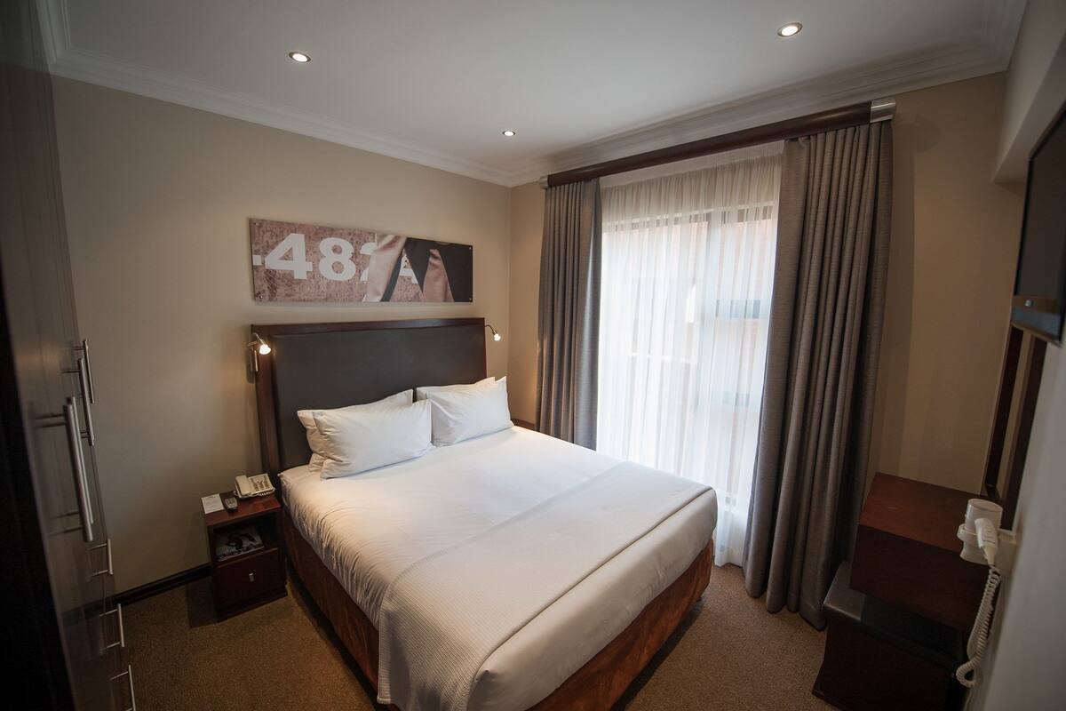 A hotel room with a large bed, a nightstand with a phone, a wall-mounted artwork, and a window with curtains.