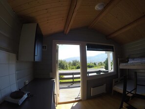 Quarto compacto com teto de madeira, janela com vista para vegetação e montanhas, uma cama beliche e pequena área de cozinha.