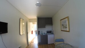 Un espacio habitable compacto con kitchenette, zona de comedor con mesa y sillas pequeñas, televisor montado y un cuadro en la pared.