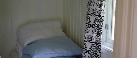 Una cama con almohadas blancas y azules, una ventana con cortina estampada y una pared blanca con líneas verticales.