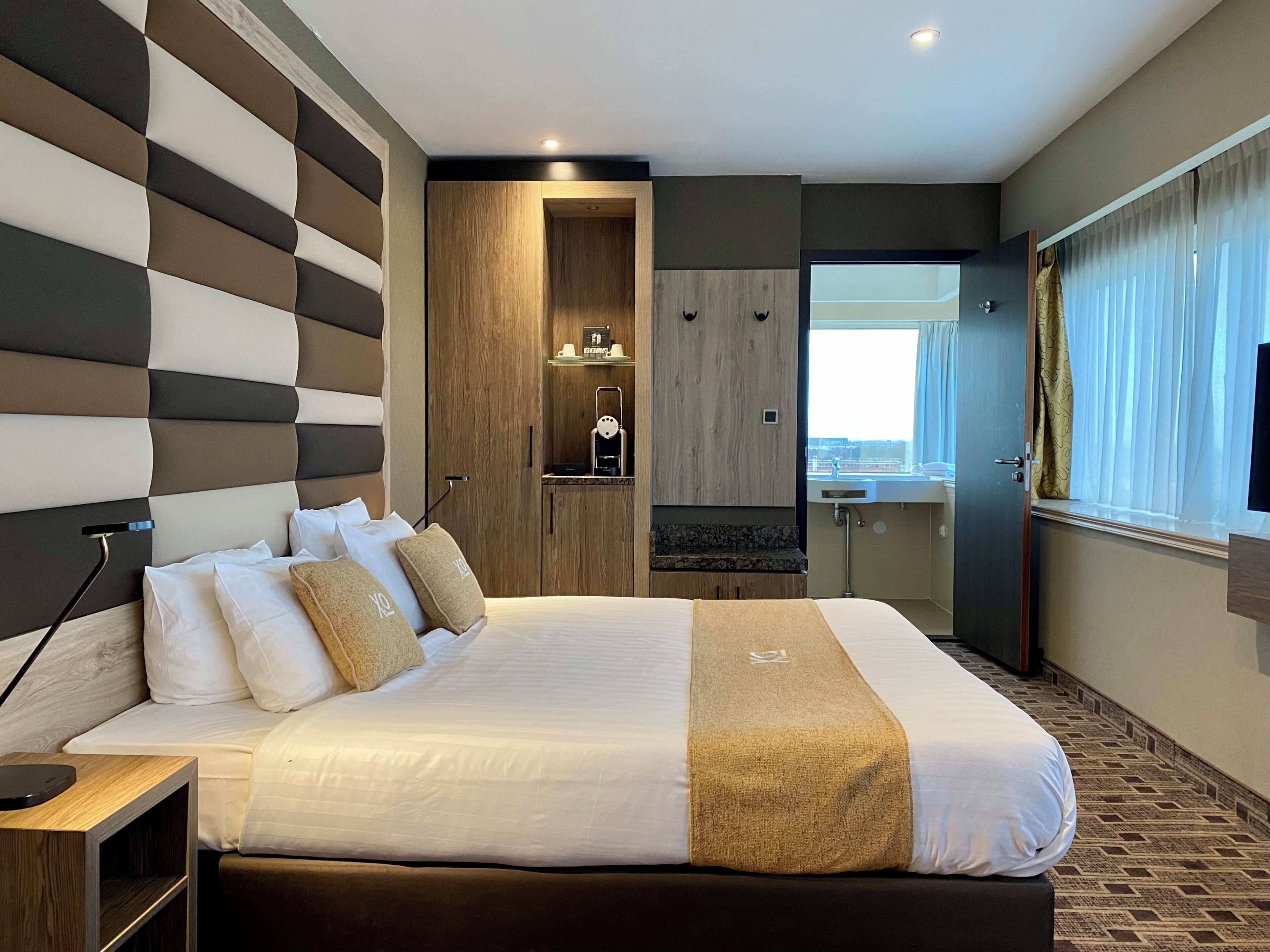 Quarto de hotel moderno com cama grande, armário de madeira, banheiro pequeno e vista para a cidade.