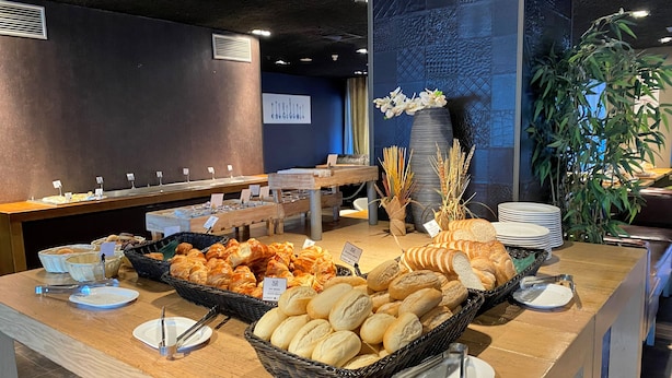 A buffet setup with various breads and pastries, a long table with multiple serving stations, and a decorative vase with flowers. - XO Hotels Blue Tower (Amsterdam)