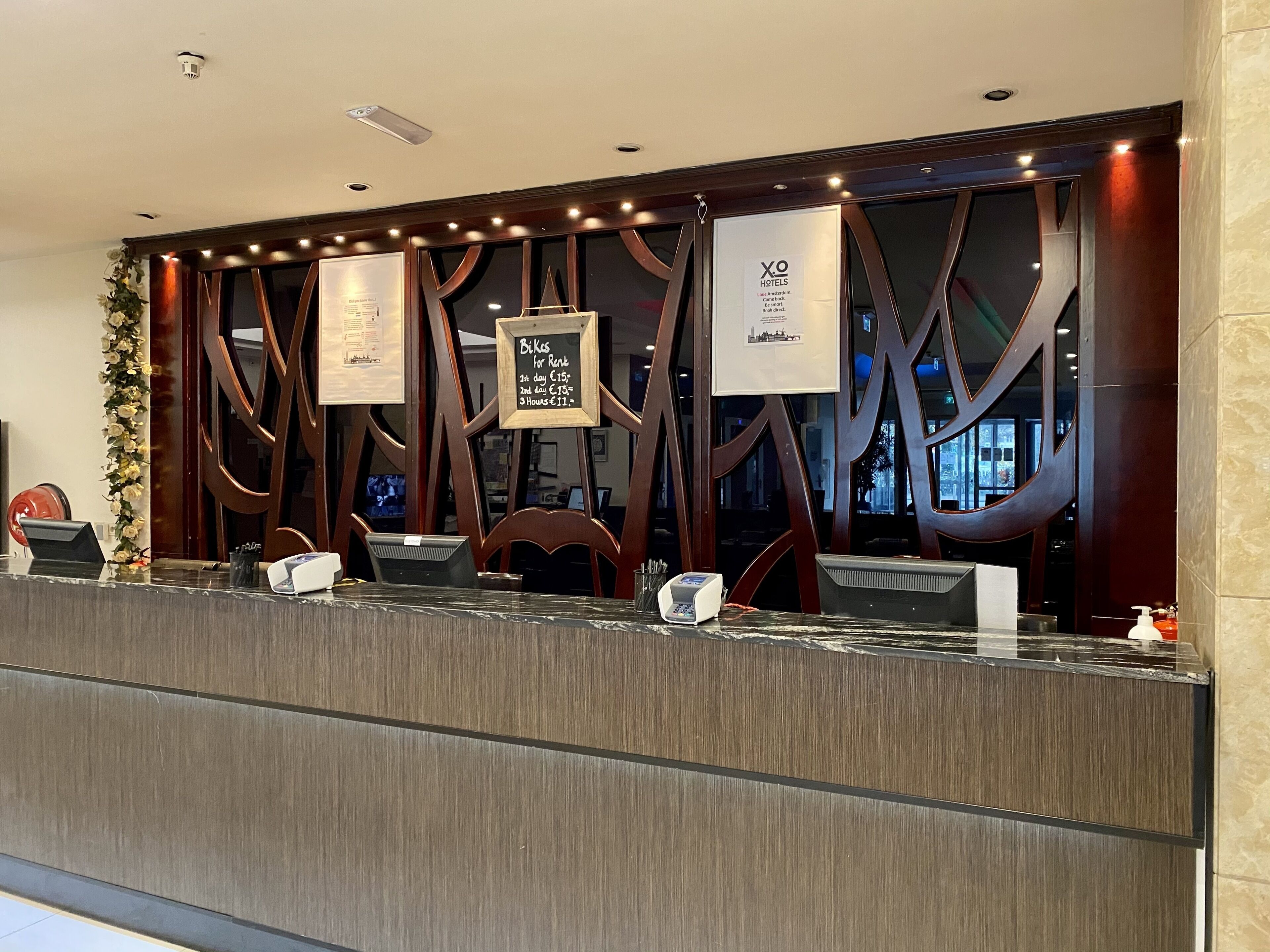 Balcão de recepção de hotel com design moderno, com um painel de parede em madeira, um letreiro emoldurado para aluguel de bicicletas e um terminal de check-in digital.