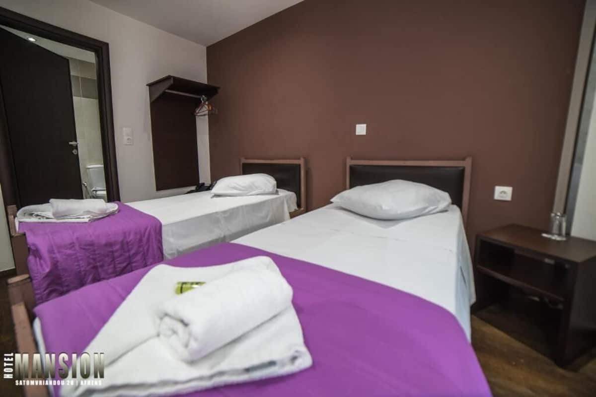 Habitación de hotel con dos camas, cada una con una almohada y una toalla doblada. Hay una mesita pequeña con una toalla encima, y una mesita de noche con una lámpara.
