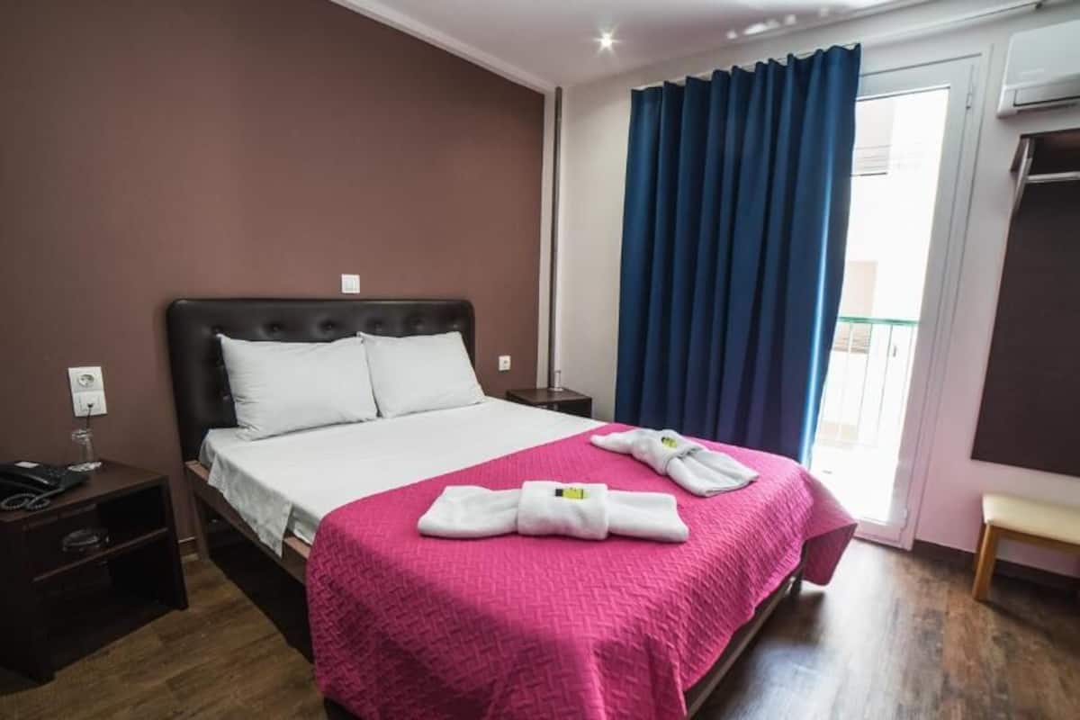 Una habitación de hotel con cama, mesita de noche, banco, armario y ventana con cortinas.