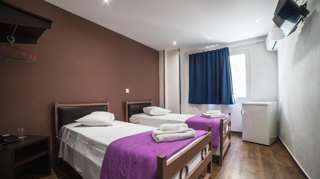Habitación de hotel con dos camas, una mesita, un calentador, una ventana con cortinas y un aire acondicionado de pared.