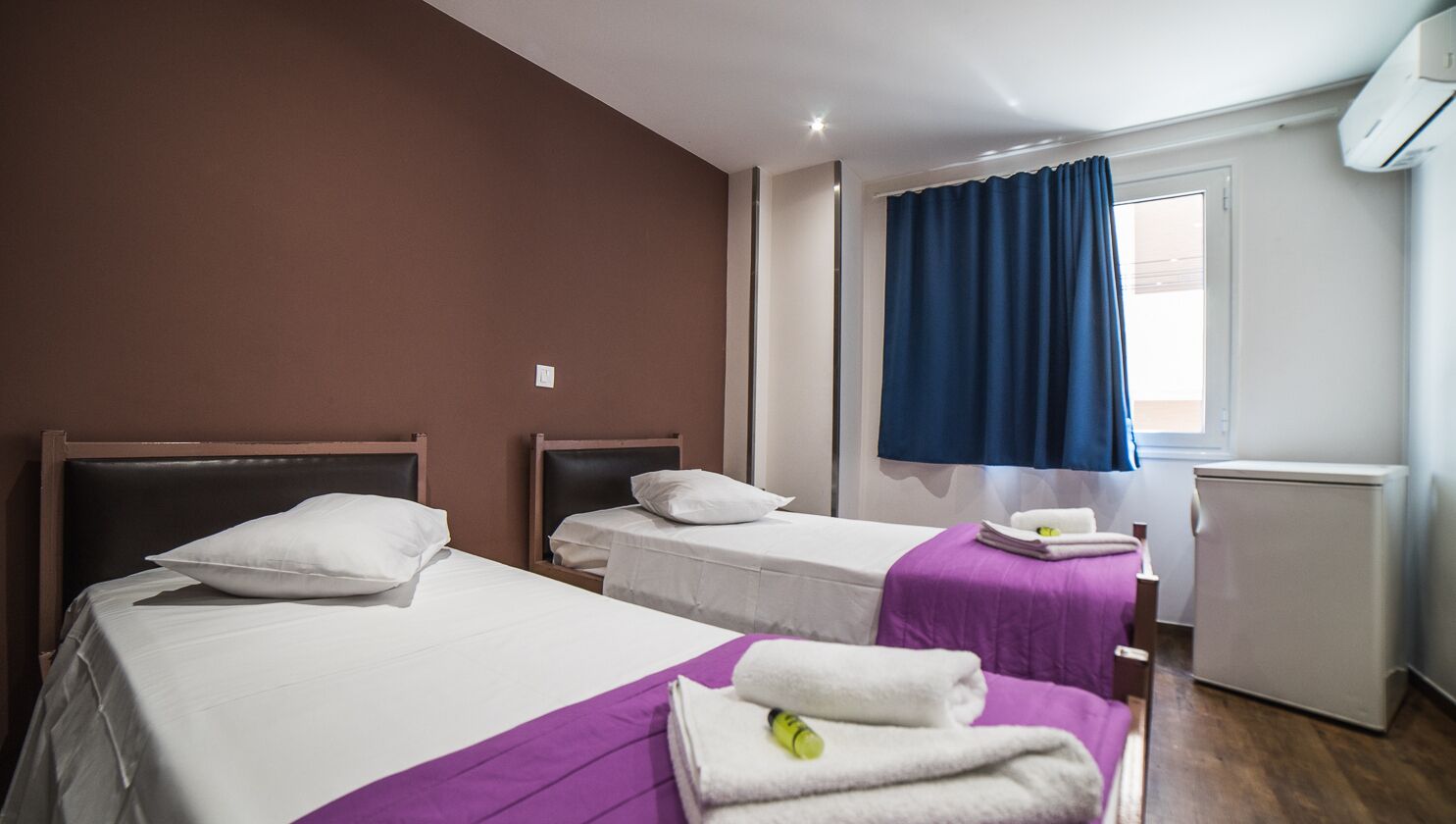 Habitación de hotel con dos camas, una ventana con cortinas azules, un radiador y una mesita con toallas.