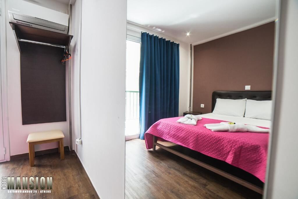 Ett hotellrum med en säng, en bänk, en garderob och ett fönster med gardiner.