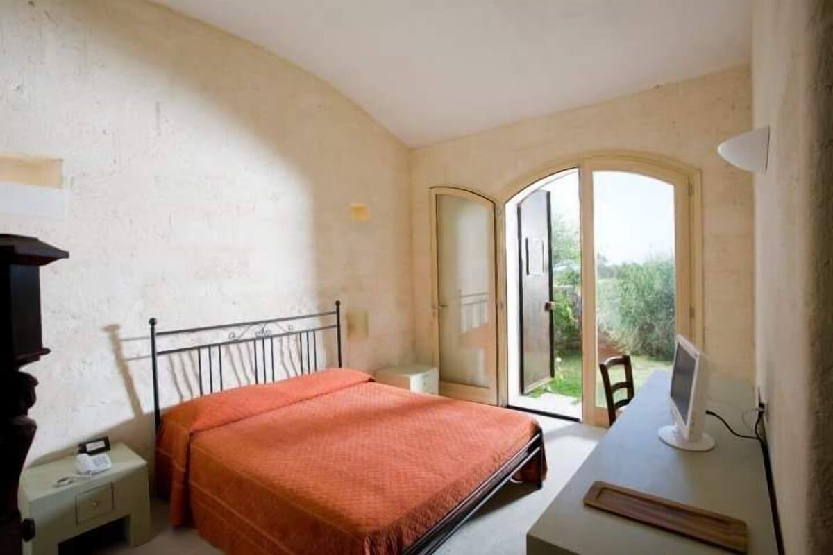 Un dormitorio con cama, escritorio con computadora, mesita de noche y puerta corrediza de vidrio que da a un balcón.