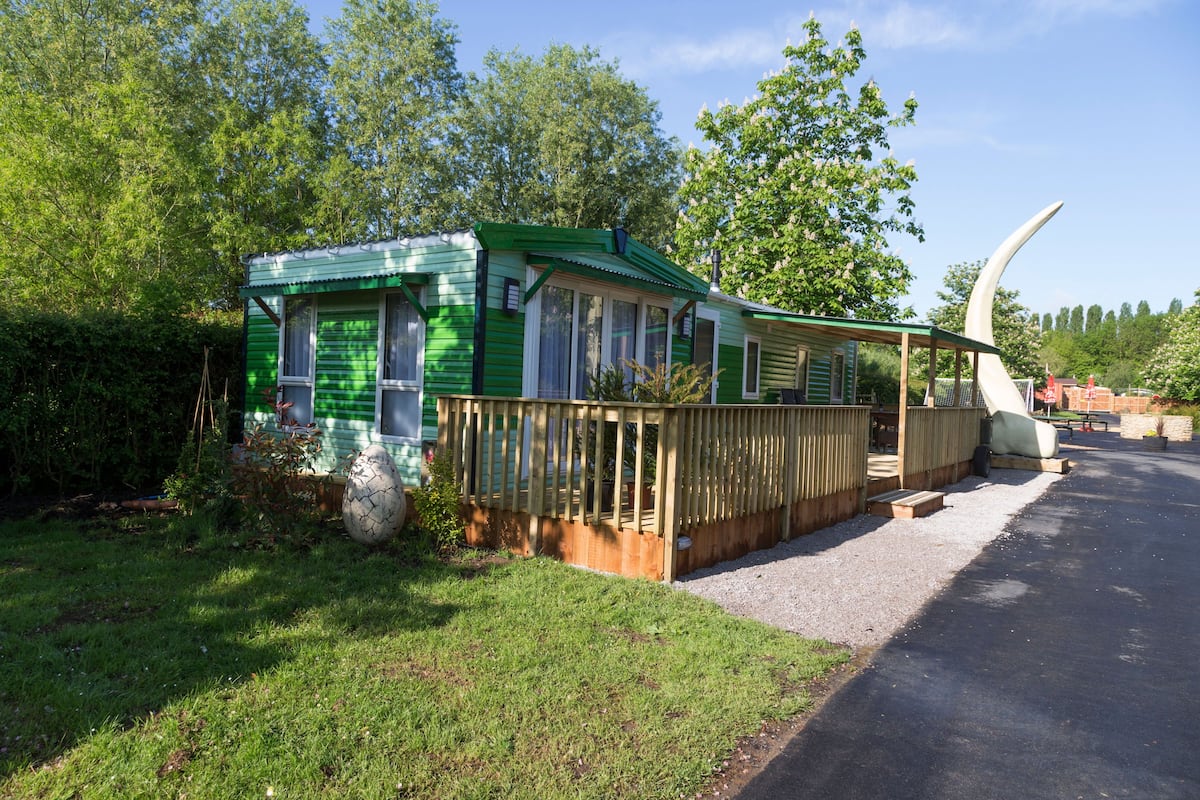 Une maison mobile verte avec une véranda clôturée, une grande sculpture blanche et une table de pique-nique à l’arrière-plan.