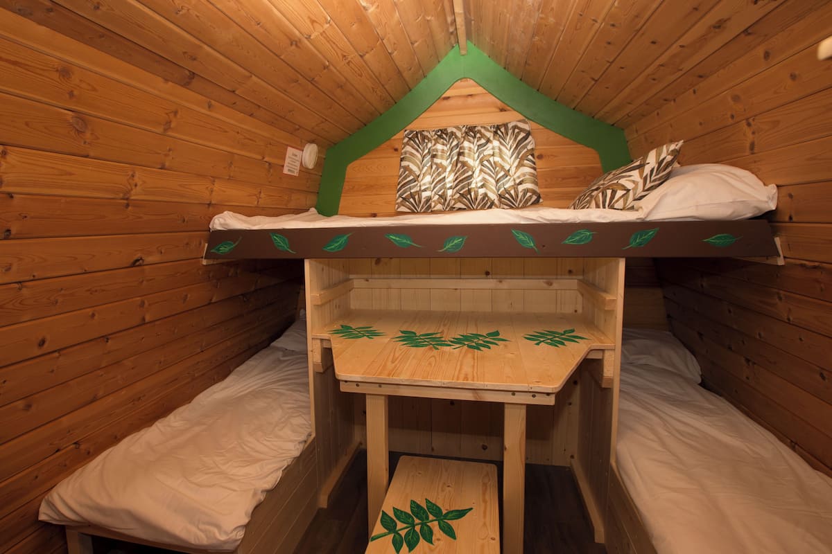 Une cabane en bois avec des lits superposés, une petite table et une tête de lit ornée d’un motif de feuilles vertes.