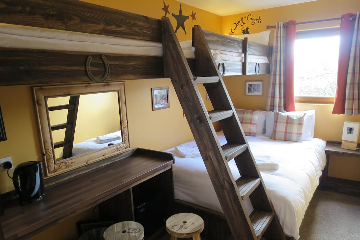 Un lit superposé avec une échelle, un bureau avec un miroir, des tabourets et un lit avec des oreillers et une couverture.