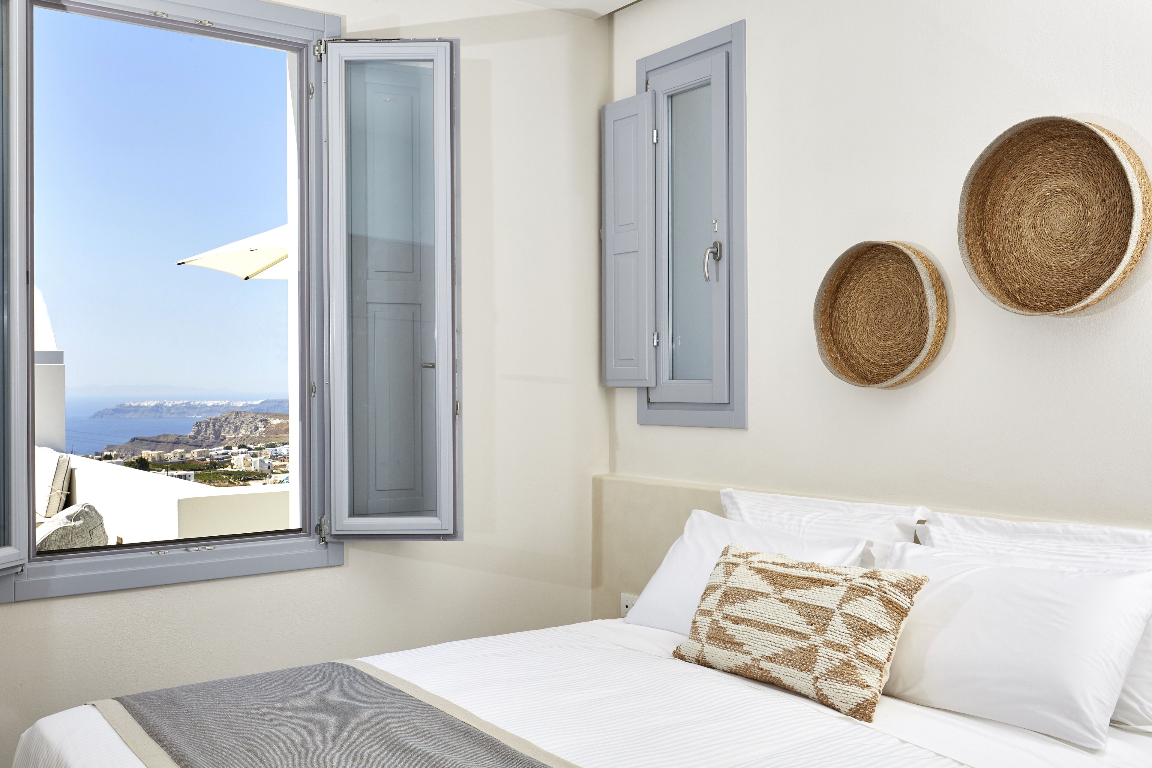 A bedroom with a bed, white bedding, a view of the sea, and two woven baskets on the wall.