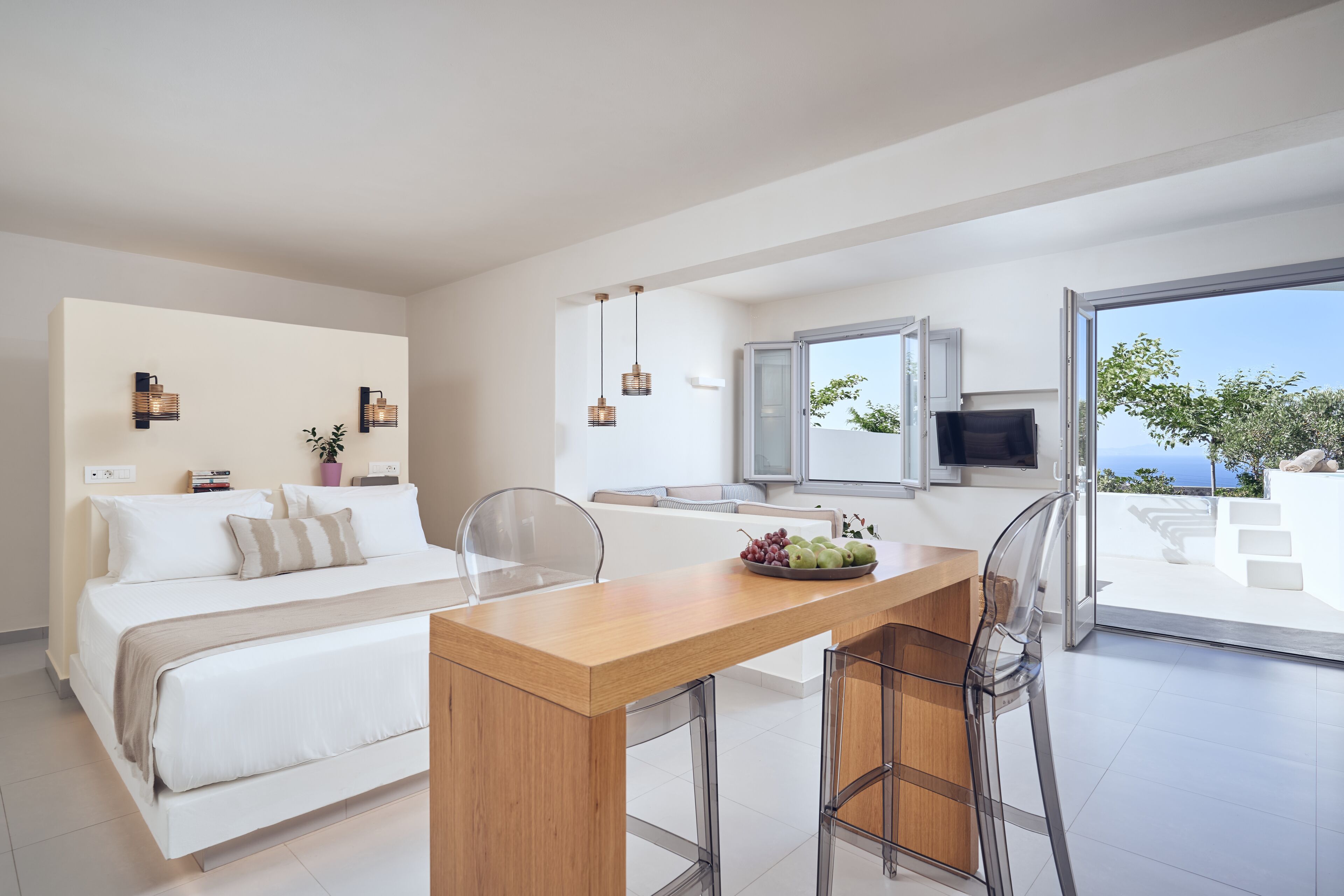 A modern hotel room with a bed, a wooden dining table with two chairs, a TV, and a balcony with a view of the sea.
