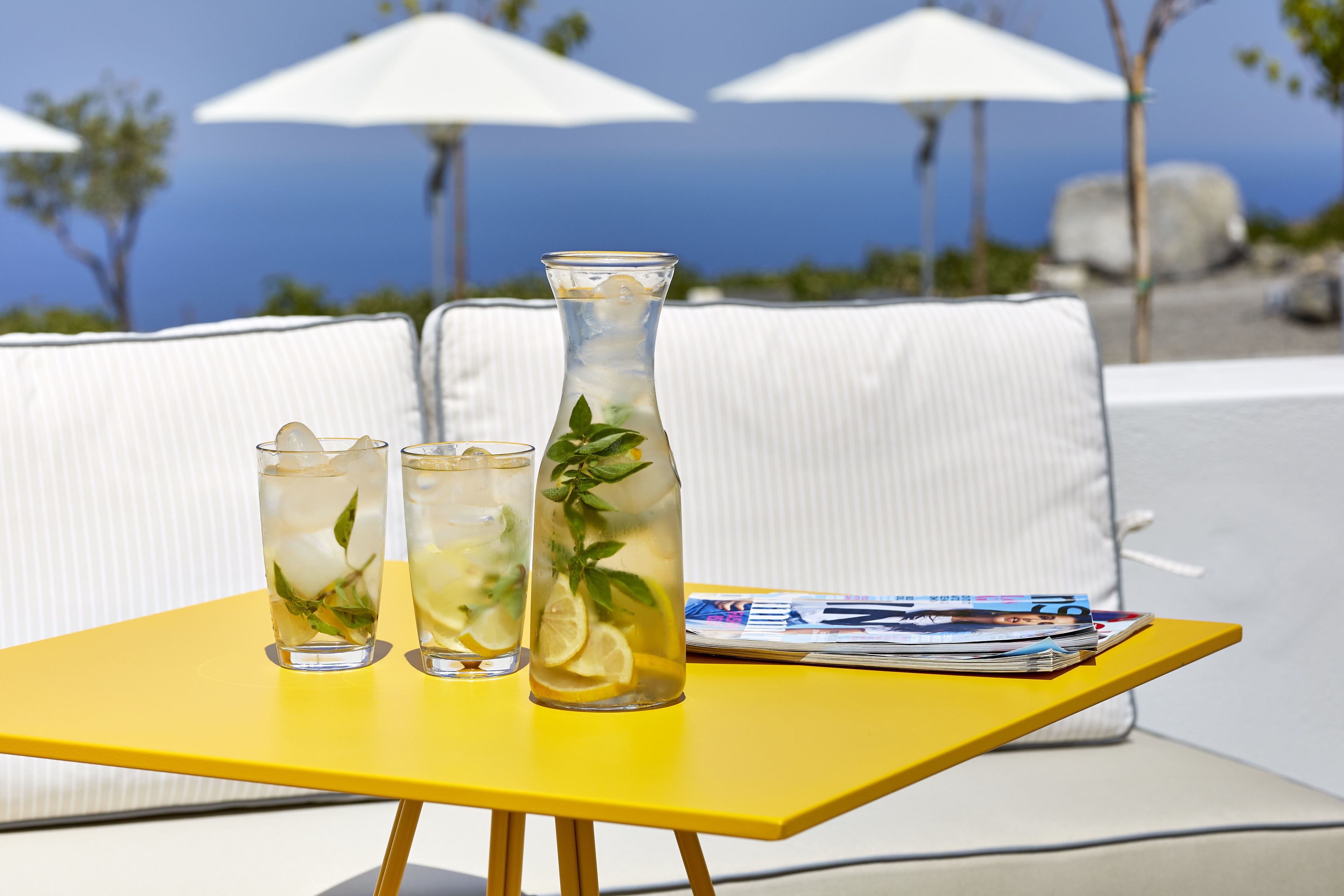 A yellow table with two glasses of iced drinks and a tall glass with a plant, set on a white sofa with magazines.