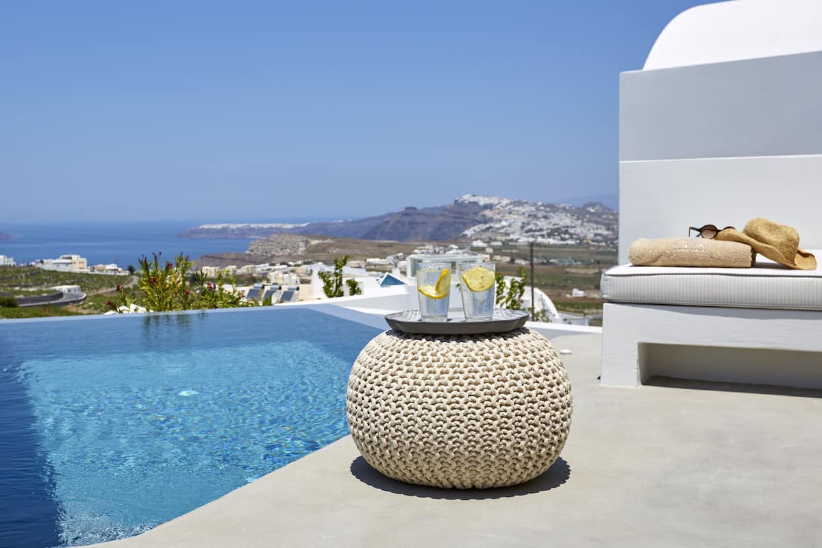 Zona junto a la piscina con vista al mar, un taburete redondo de fibras naturales y un banco blanco con cojín, toallas y gafas de sol.