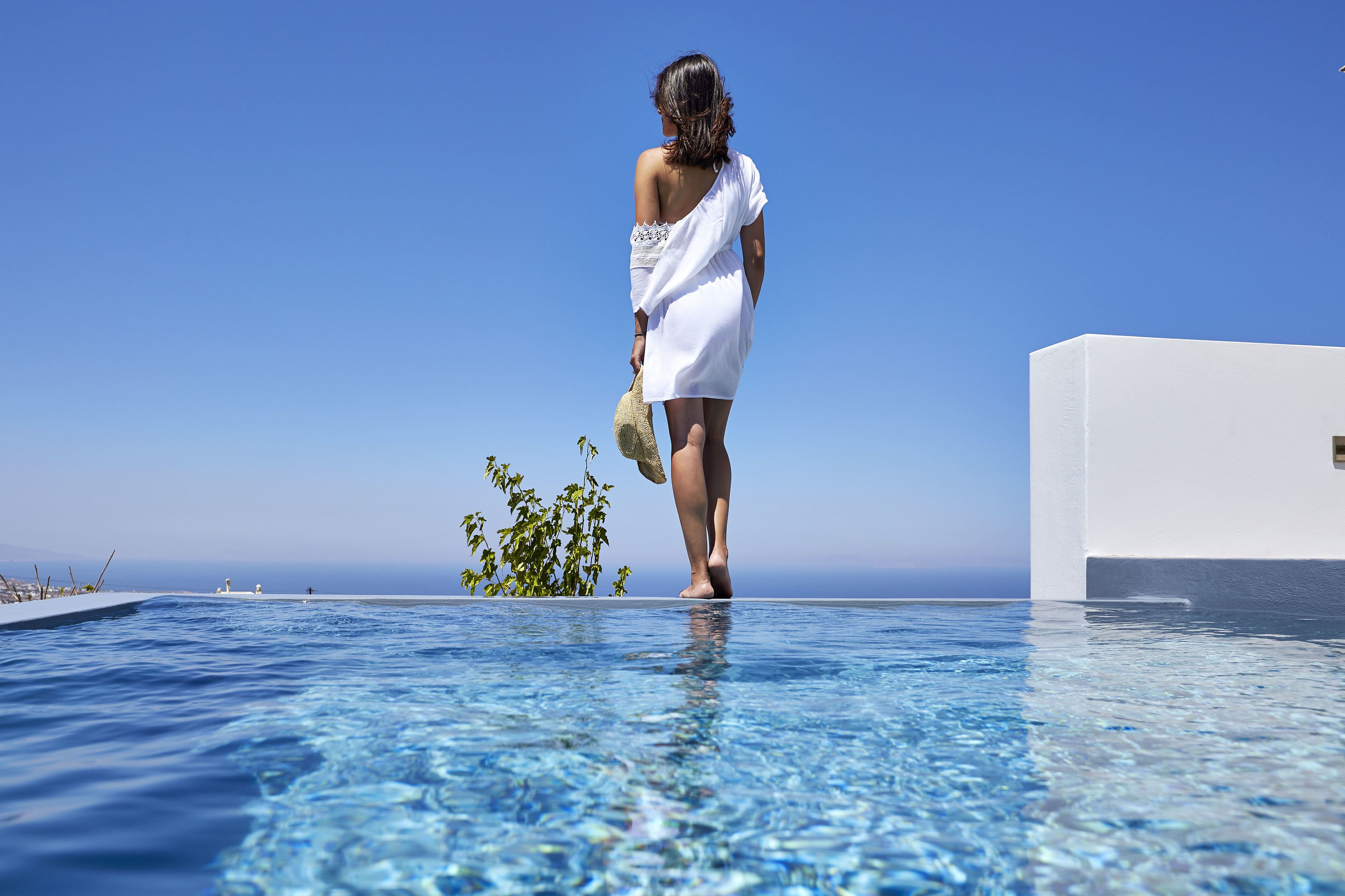 A woman in a white dress walking on a poolside with a clear view of the ocean and a modern building in the background.