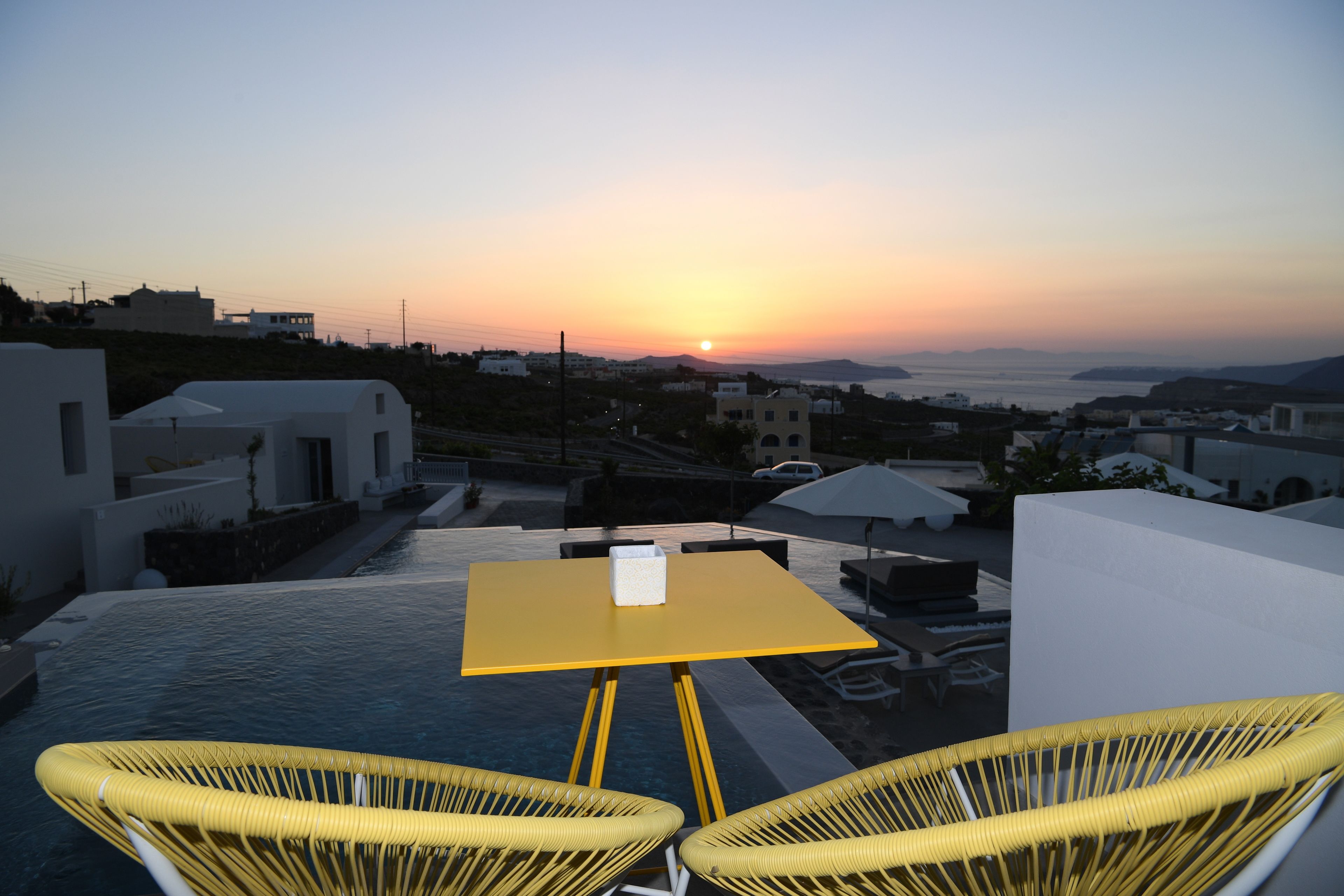Una terraza con una mesa amarilla y dos sillas de mimbre, con vistas a un atardecer sobre un paisaje costero.