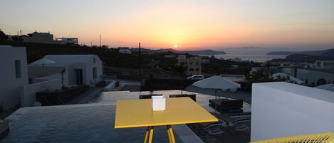 Una terraza con una mesa amarilla y dos sillas de mimbre, con vistas a un atardecer sobre un paisaje costero.