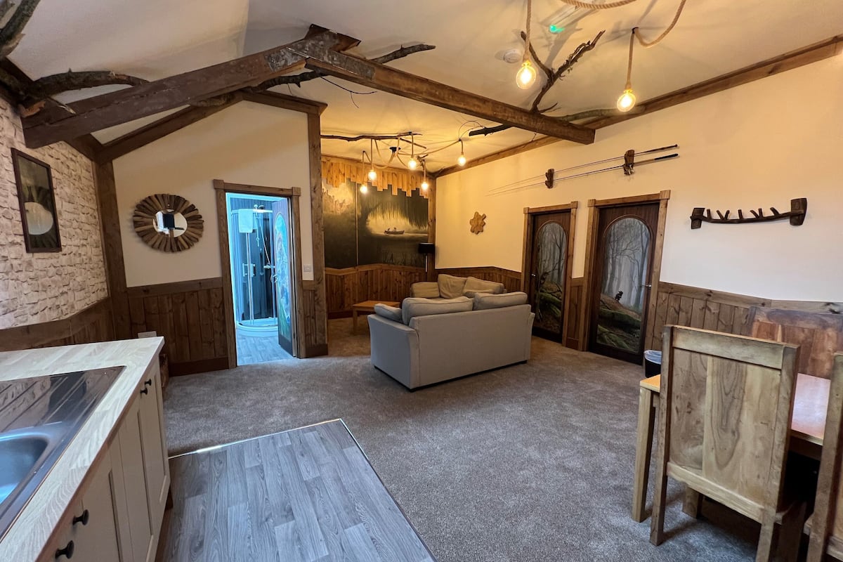 A rustic interior with exposed wooden beams, a stone wall, a modern kitchen, a seating area with a sofa, and a shower room.