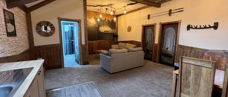 Un interior rústico con vigas de madera a la vista, una pared de piedra, una cocina moderna, una zona de estar con sofá y un cuarto de baño con ducha.