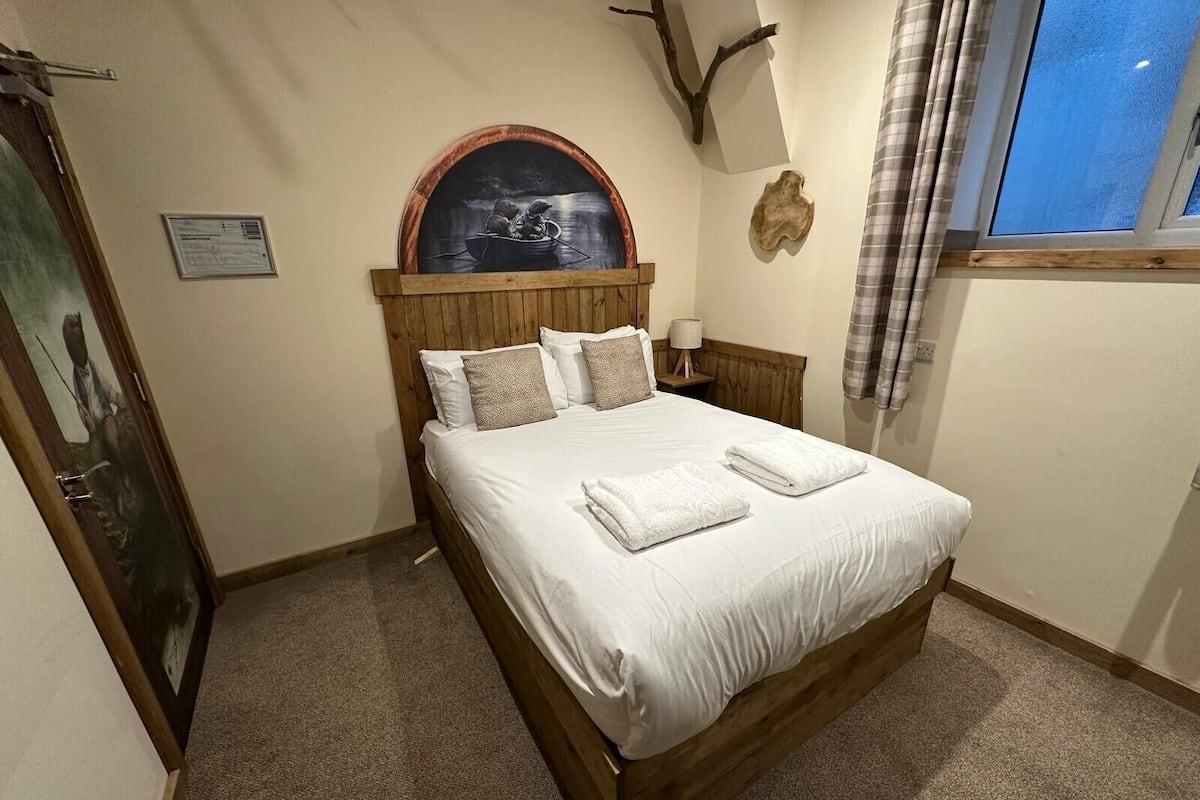 A bedroom with a wooden bed, a nightstand, a wall-mounted lamp, a framed picture, and a window with curtains.