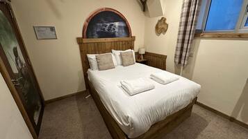 A bedroom with a wooden bed, a nightstand, a wall-mounted lamp, a framed picture, and a window with curtains.