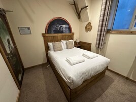 A bedroom with a wooden bed, a nightstand, a wall-mounted lamp, a framed picture, and a window with curtains.