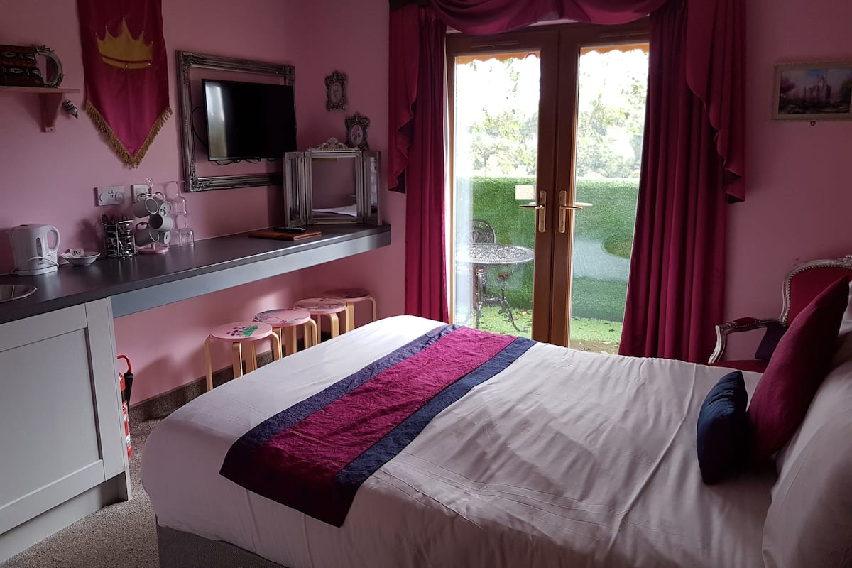 A bedroom with a bed, a desk with a mirror, a television, and a view of a garden through a sliding door.