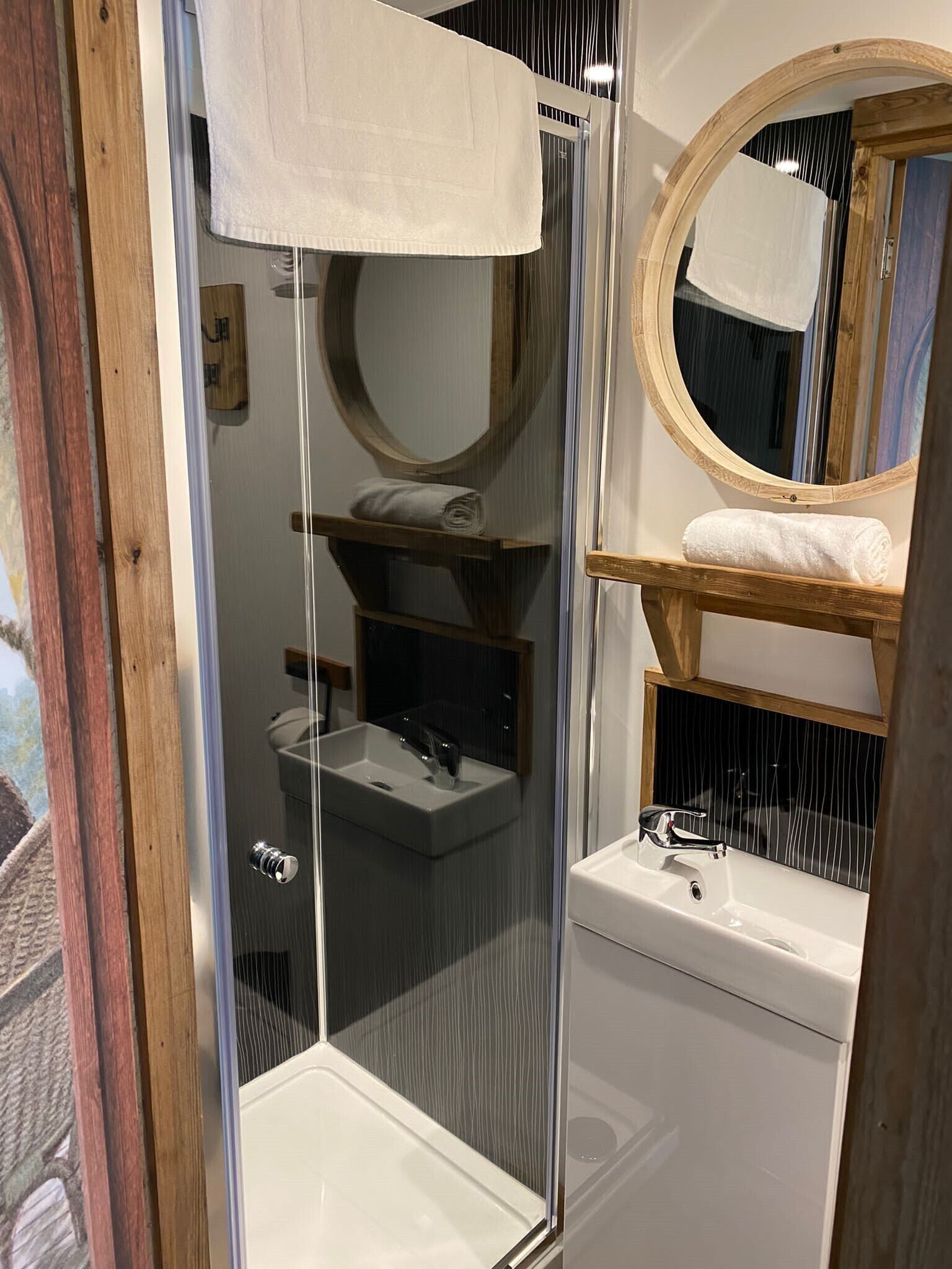 Un bany amb plat de dutxa de vidre, un lavabo blanc i una prateleira de fusta amb una tovallola.