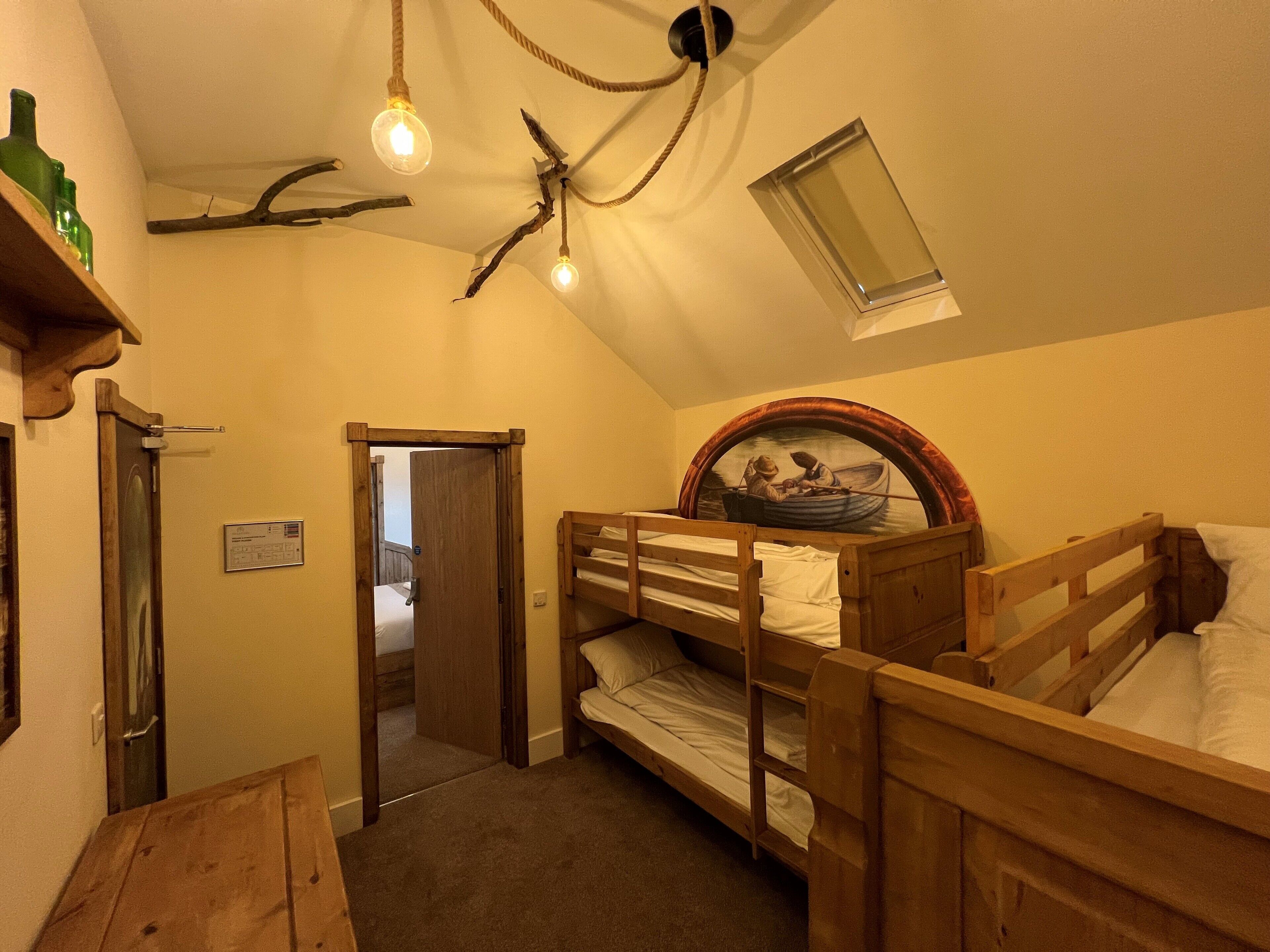 Et sovesal med køjesenge, en loftlampe, et lille vindue og en dør til et andet rum.