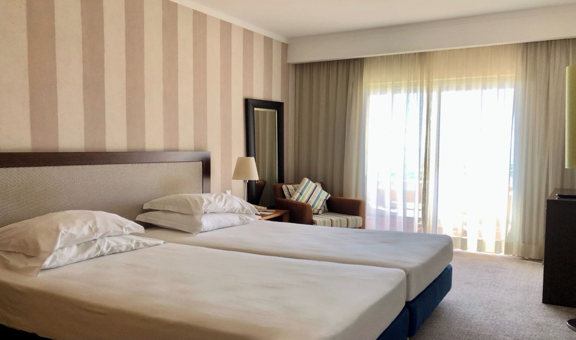 Hotellihuone, jossa on kaksi sänkyä, yöpöytä, lamppu, tuoli ja suuri ikkuna, jossa on verhot.