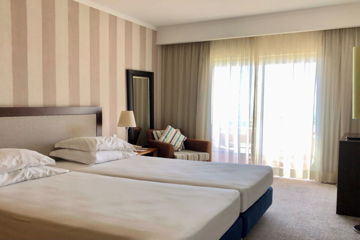 Ein Hotelzimmer mit zwei Betten, einem Nachttisch, einer Lampe, einem Sessel und einem großen Fenster mit Vorhängen.