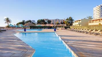 Piscina con sillas de descanso y sombrillas, rodeada de edificios y palmeras.