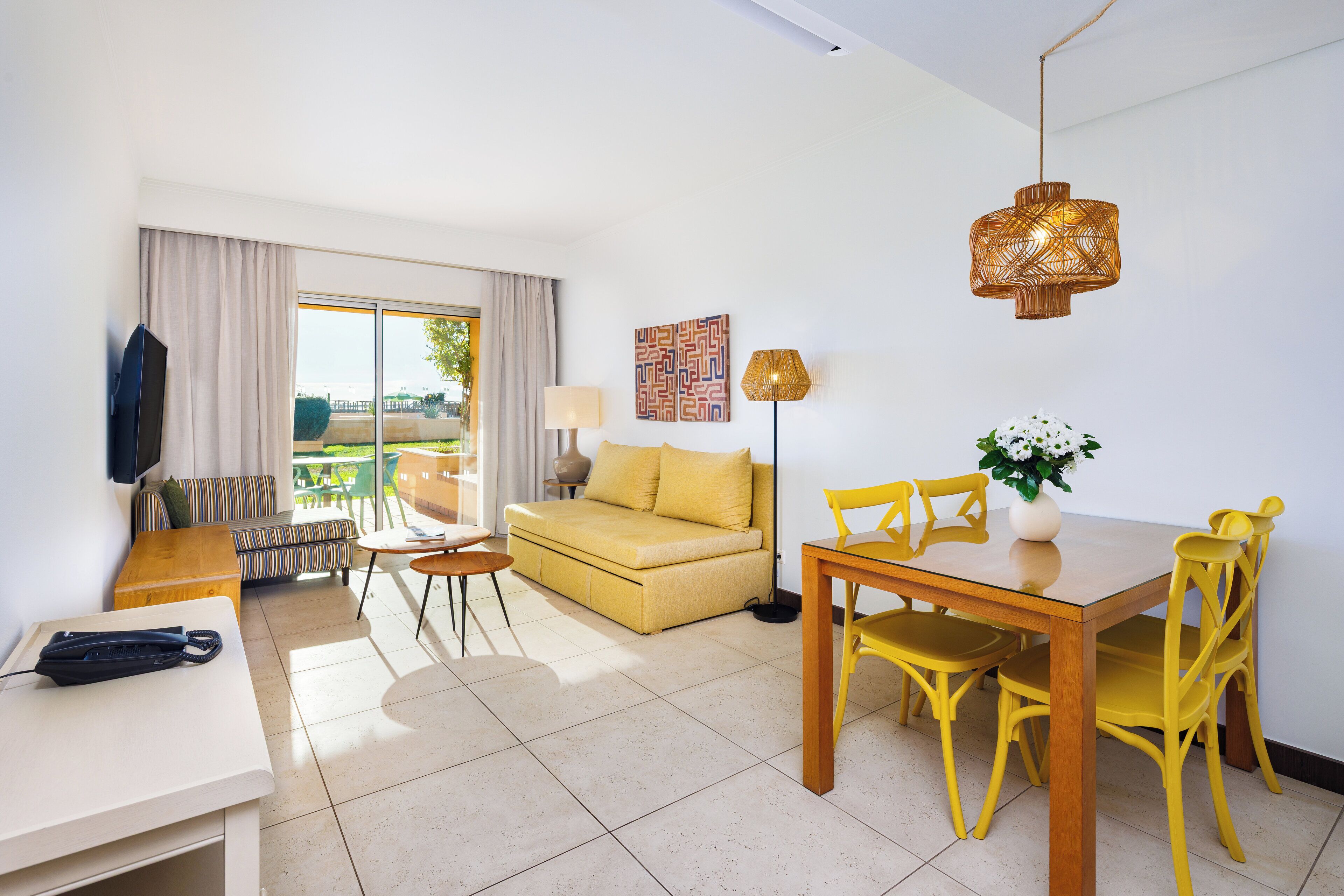 A modern living room with a sofa, dining table, and chairs, a flat-screen TV, and a large window with curtains.