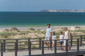 A family walking on a wooden boardwalk by the beach with cliffs in the background. - Pestana Alvor Beach Villas (Portimão)